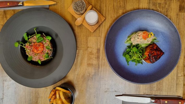 Gourmet dishes on a wooden table with cutlery and a small bowl of chips, offering a fine dining experience