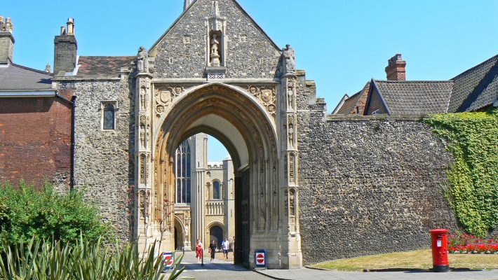 Historic stone gate entrance to Norwich Cathedral under a clear blue sky with surrounding greenery and vibrant flowers