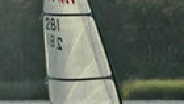 Sailboat gliding on a serene lake with lush trees in the background under a cloudy sky