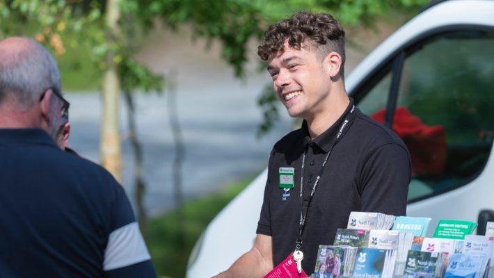 Man smiling outdoors with brochures and speaking to person in casual setting, wearing a name badge and lanyard