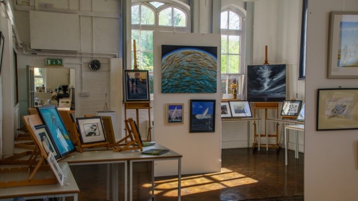 Art gallery interior with various paintings displayed on easels and walls in a well-lit room with large windows