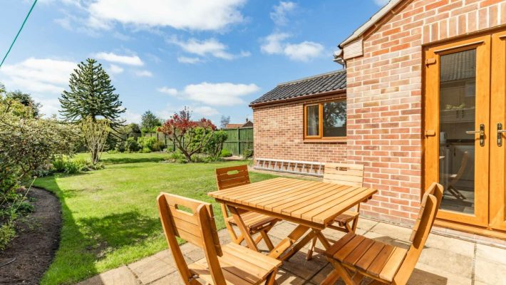 Wooden outdoor dining set on a sunny patio beside a brick house with a lush green garden in the background